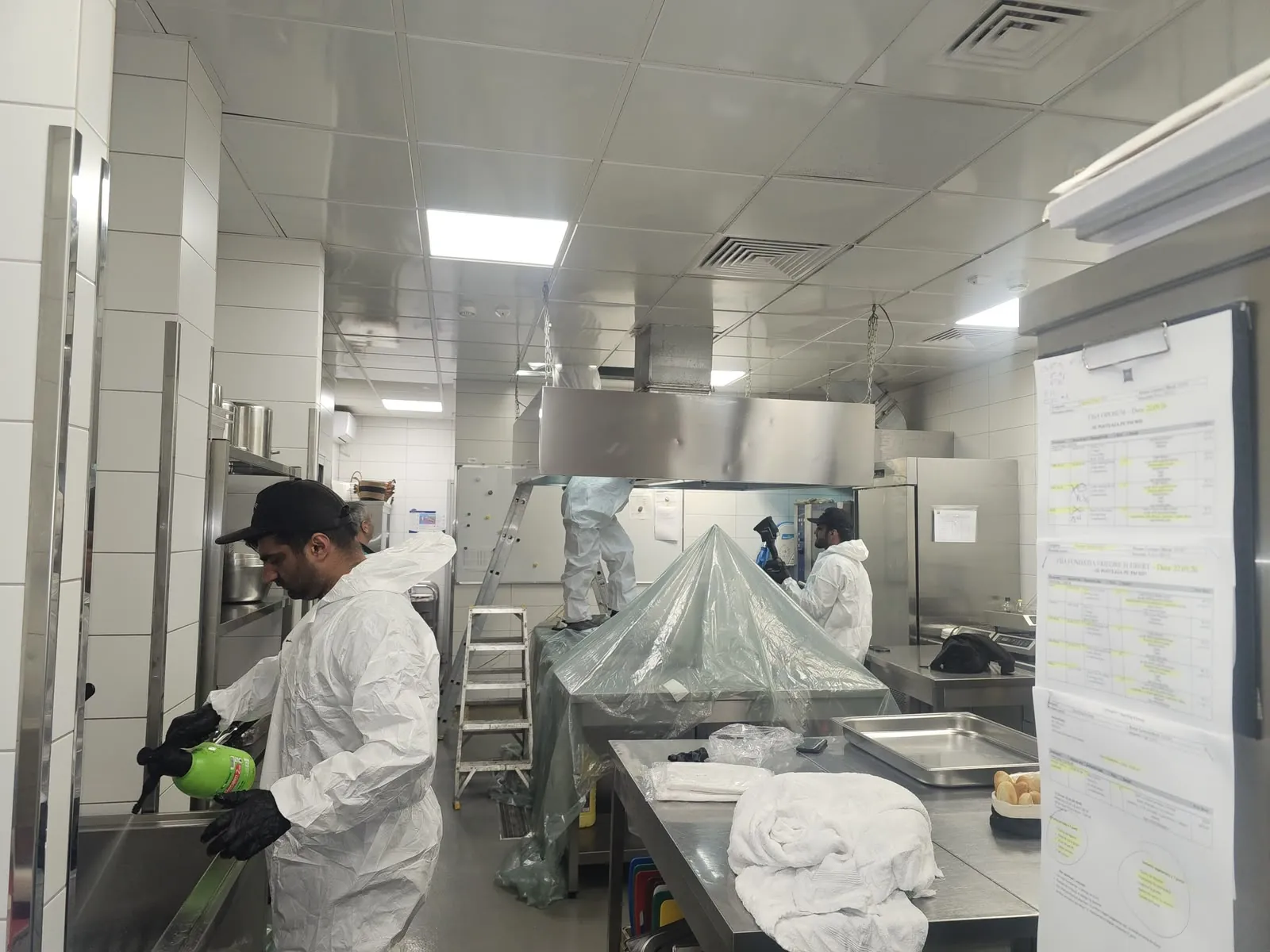 Echipă în combinezoane de protecție degresând o hotă centrală, cu utilajele bucătăriei acoperite cu folie