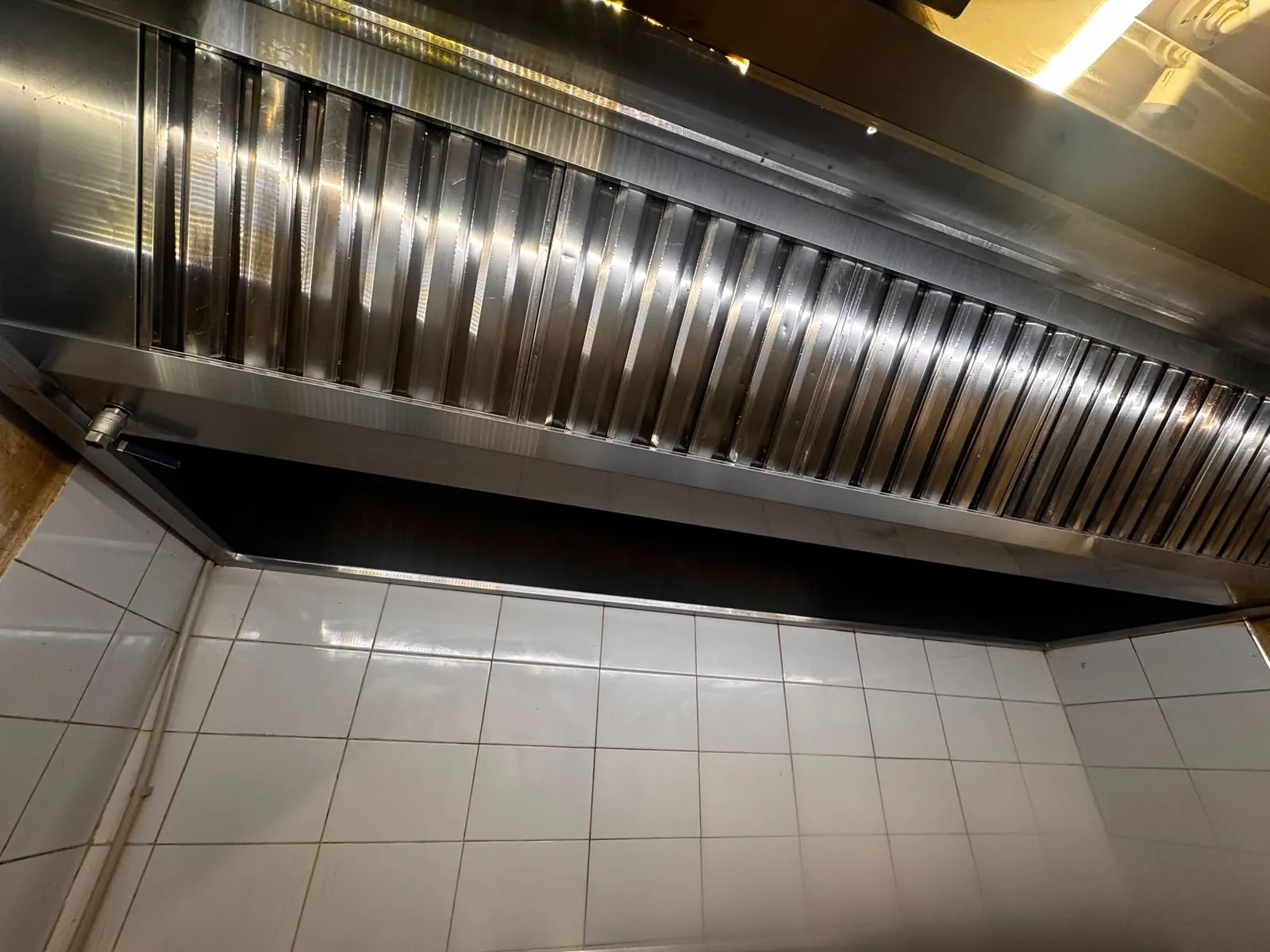 Filtre labirint din inox curate pe toată lungimea hotei, montate deasupra unui perete cu faianță albă