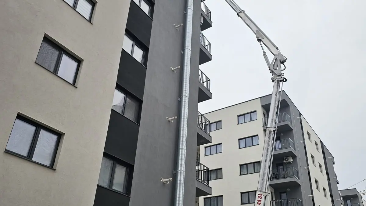 Nacelă ridicată lângă tubulatura spiralată de evacuare montată pe fațada unei clădiri cu mai multe etaje
