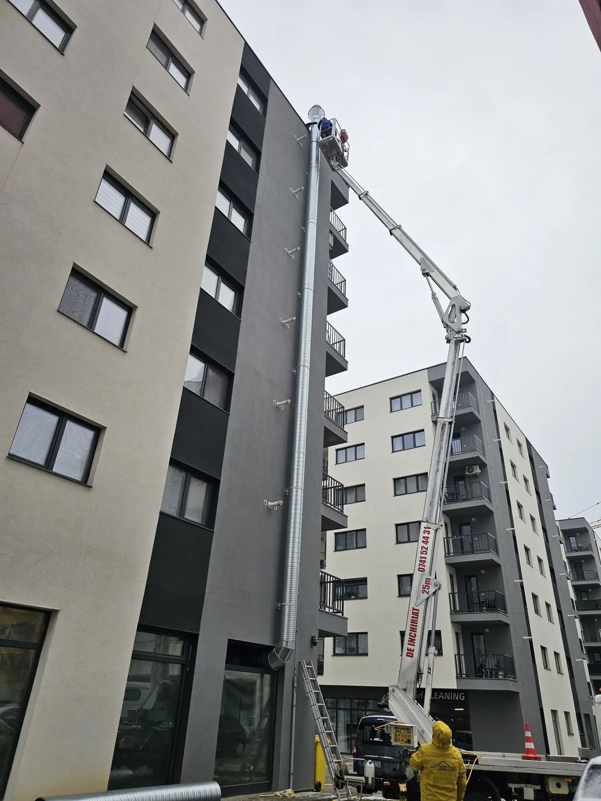Nacelă ridicată lângă tubulatura spiralată de evacuare de pe fațada unei clădiri cu mai multe etaje