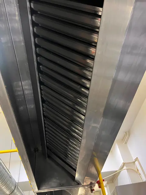Perspectivă de jos asupra filtrelor labirint curate dintr-o hotă din inox, lângă conducta de gaz galbenă
