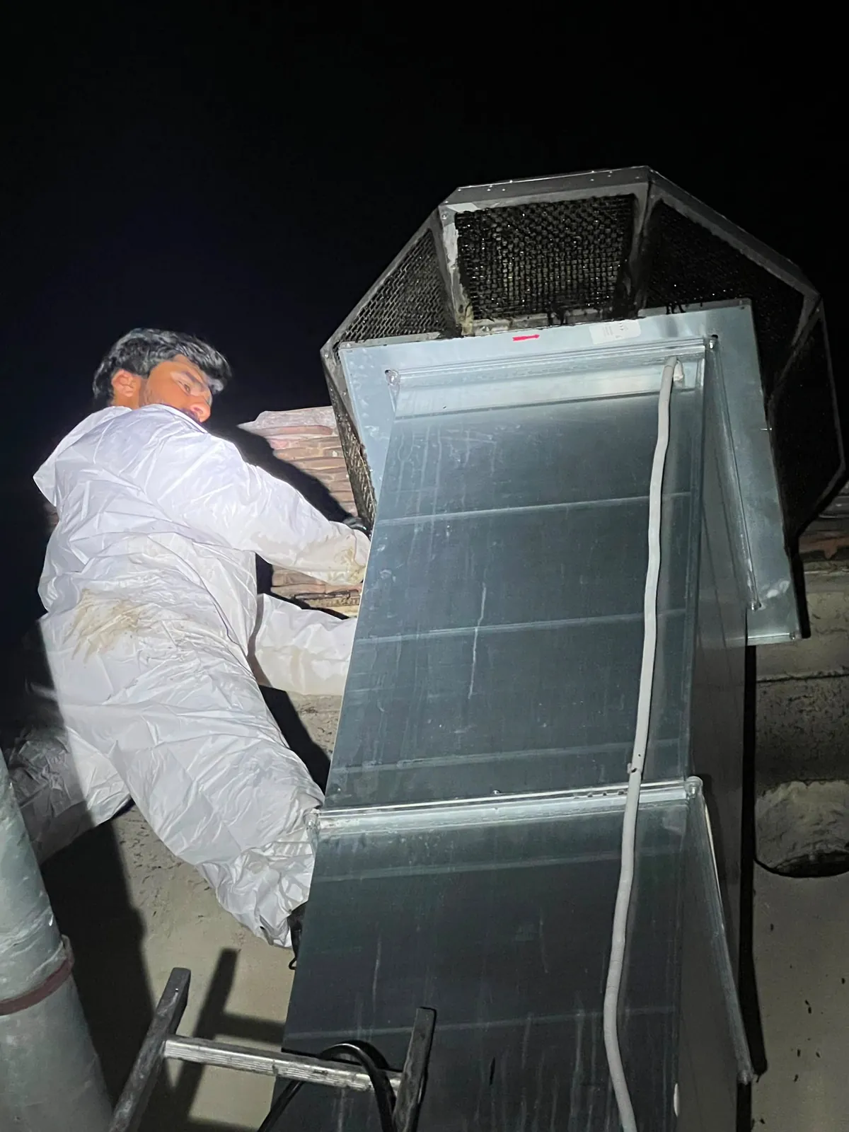 Tehnician lucrând noaptea pe acoperiș, la tubulatura rectangulară și la ventilatorul de exhaustare