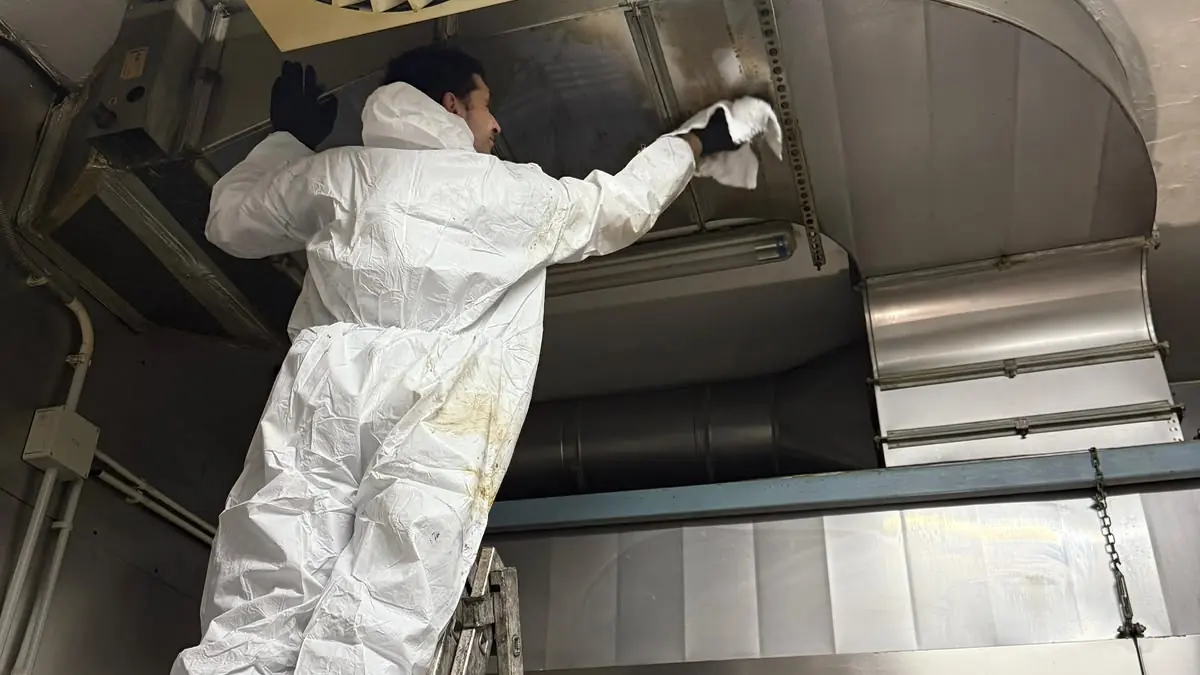 Tehnician în combinezon de protecție pe scară, ștergând tubulatura de inox de deasupra hotei după degresare