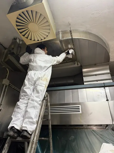 Tehnician în combinezon de protecție pe scară, ștergând tubulatura de inox de deasupra hotei după degresare