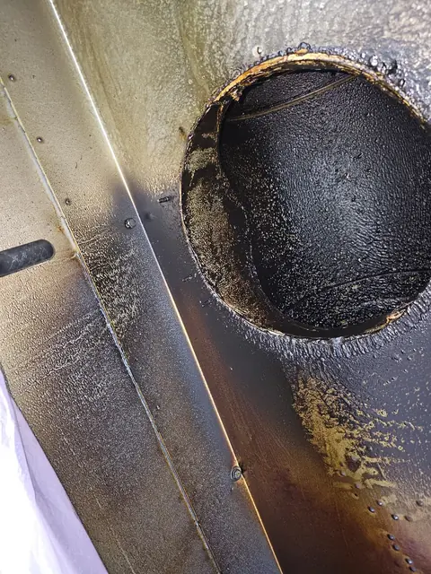 Gură de evacuare rotundă în plafonul hotei, cu crustă groasă de grăsime neagră în jurul tubulaturii, înainte de curățare